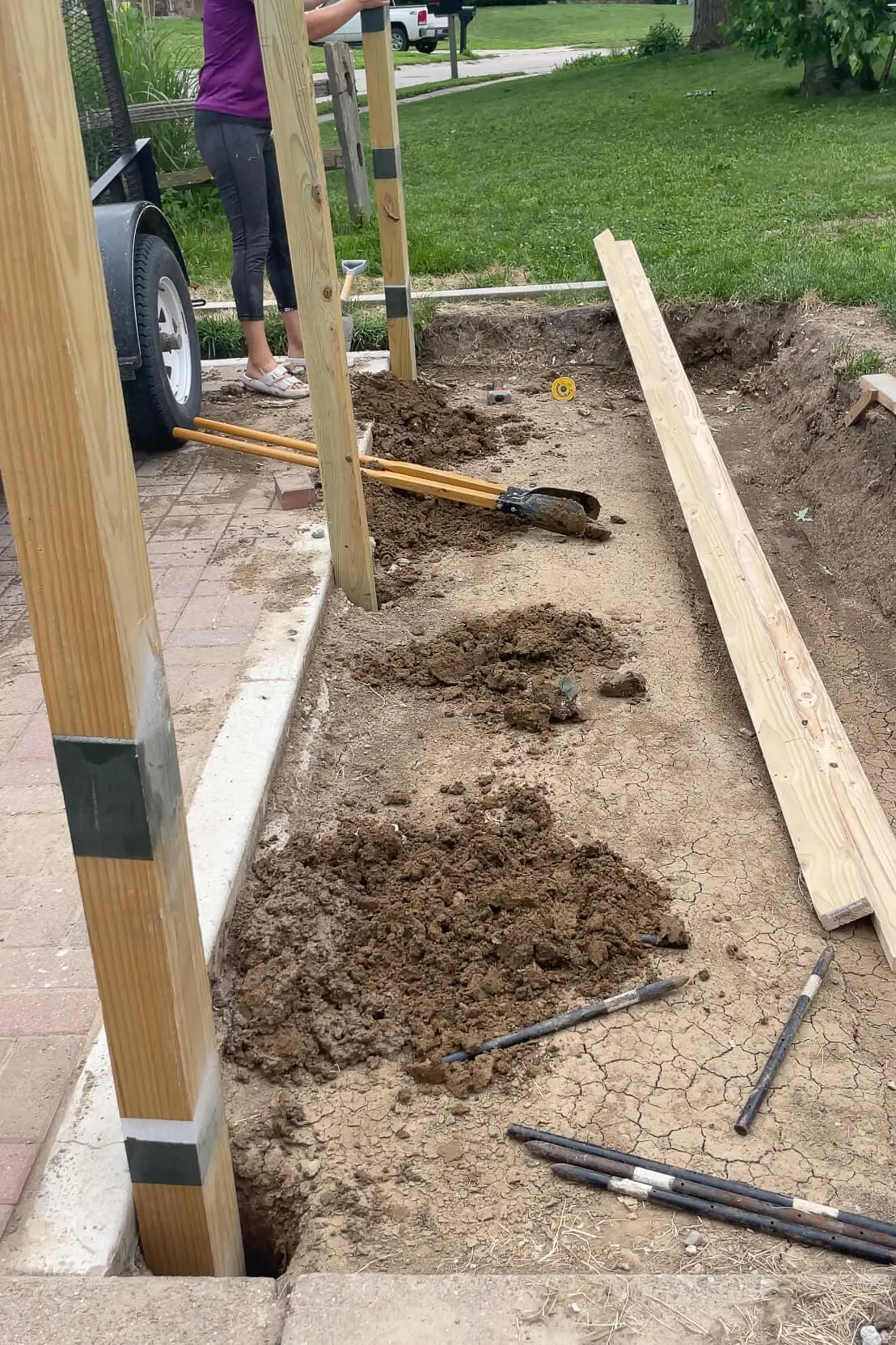 DIYer preparing post holes with a post hole digger to set wooden fence posts for a trash can enclosure project on a dirt and brick base.